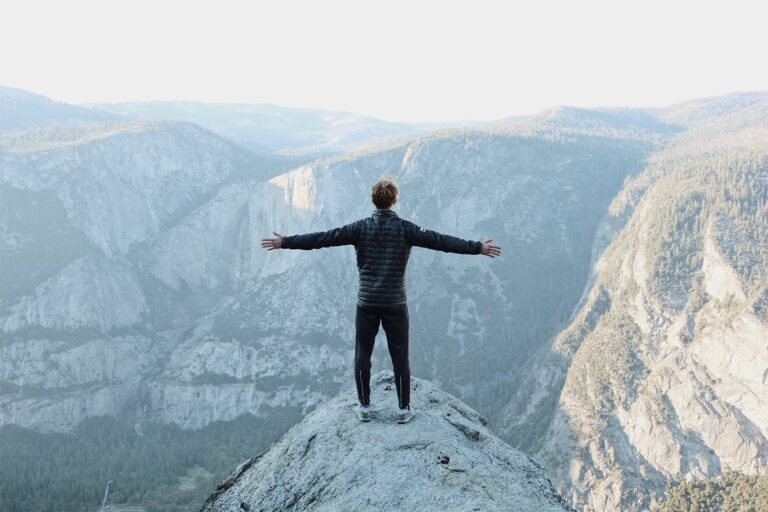 Person in a black jacket stands with arms outstretched on a rocky cliff, overlooking a vast mountain landscape.