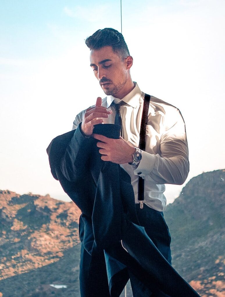 A man in a white shirt and suspenders holds a dark jacket, with a mountainous landscape visible in the background through a large window.