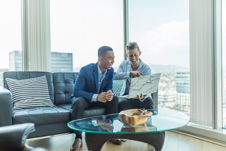Two men sitting on a black sofa in a modern office, looking at a laptop together, with a glass coffee table and cityscape visible through large windows.
