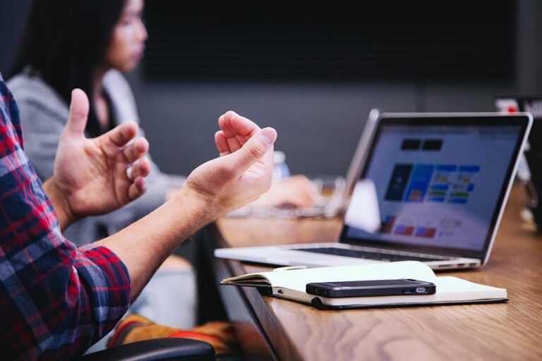 Person gesturing while sitting at a wooden table with a laptop and smartphone; another person blurred in the background.