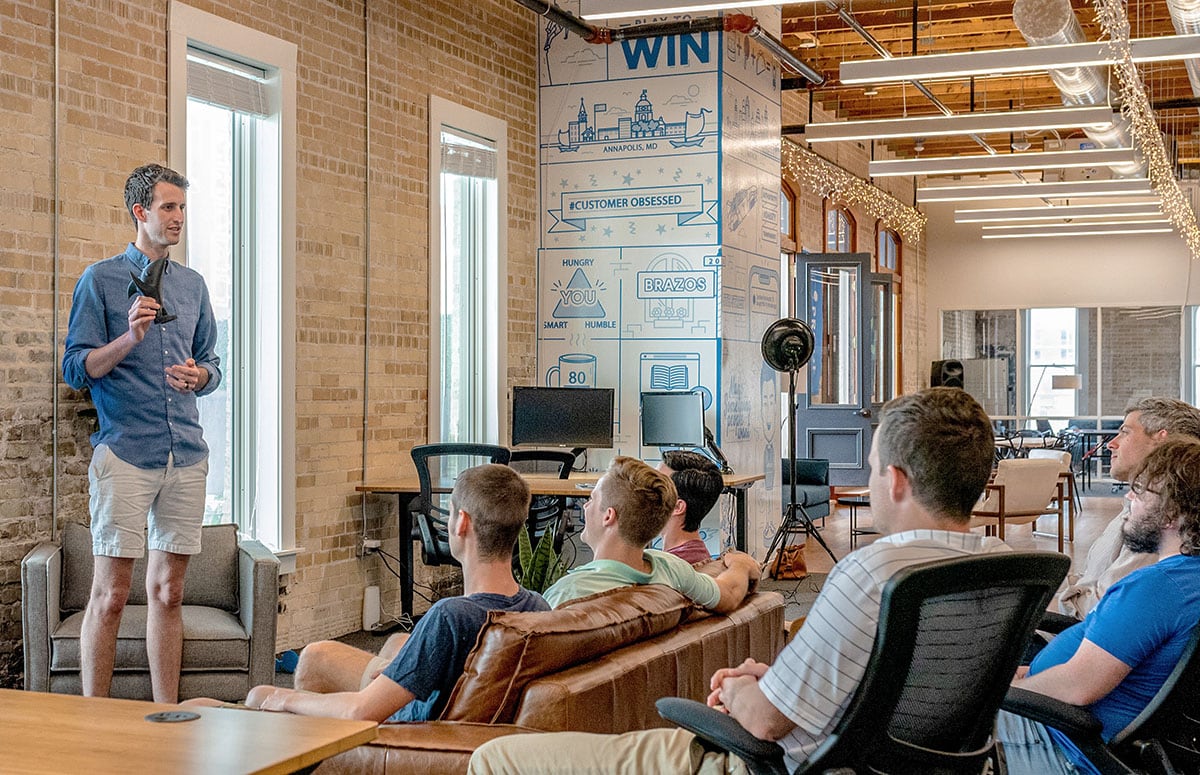 A person in a blue shirt presents to a group seated on couches in a modern office space, with exposed brick walls and various equipment.